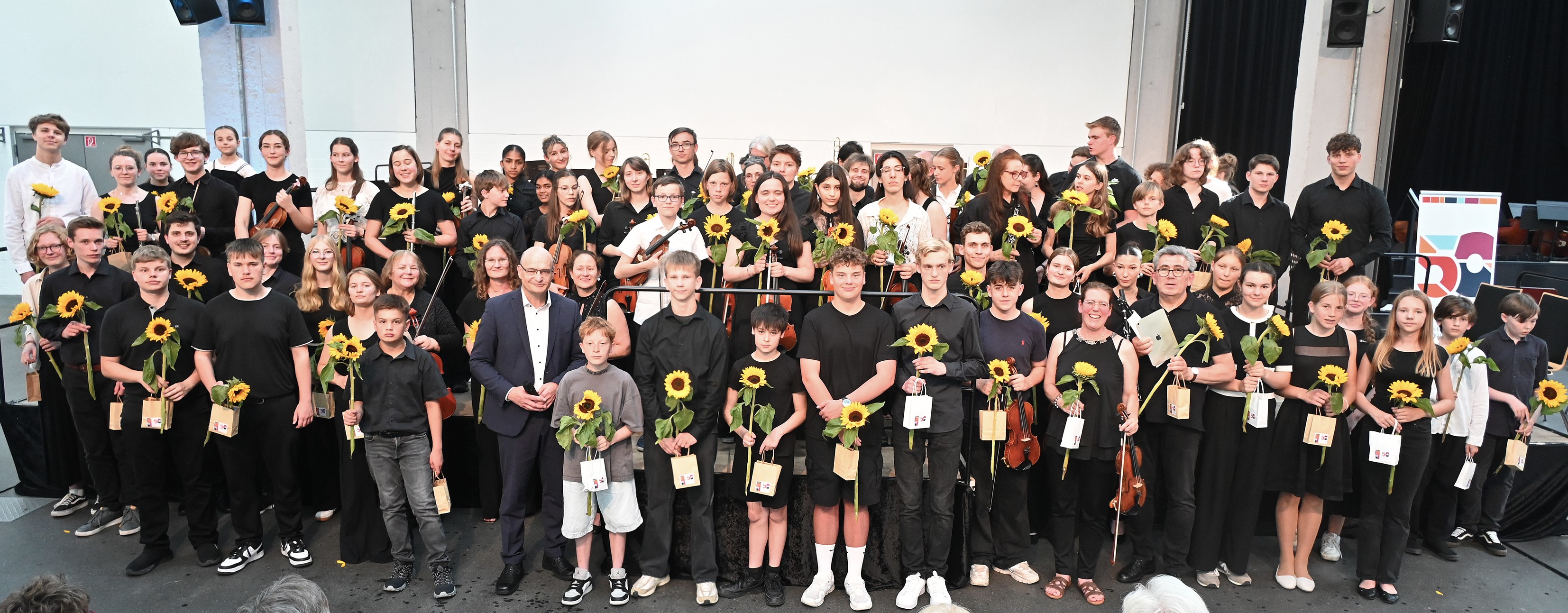 Die Musizierenden stehen auf der Bühne und davor und haben Sonnenblumen und Geschenke für ihren Auftritt bekommen. Die Musizierenden stehen auf der Bühne und davor und haben Sonnenblumen und Geschenke für ihren Auftritt bekommen.
