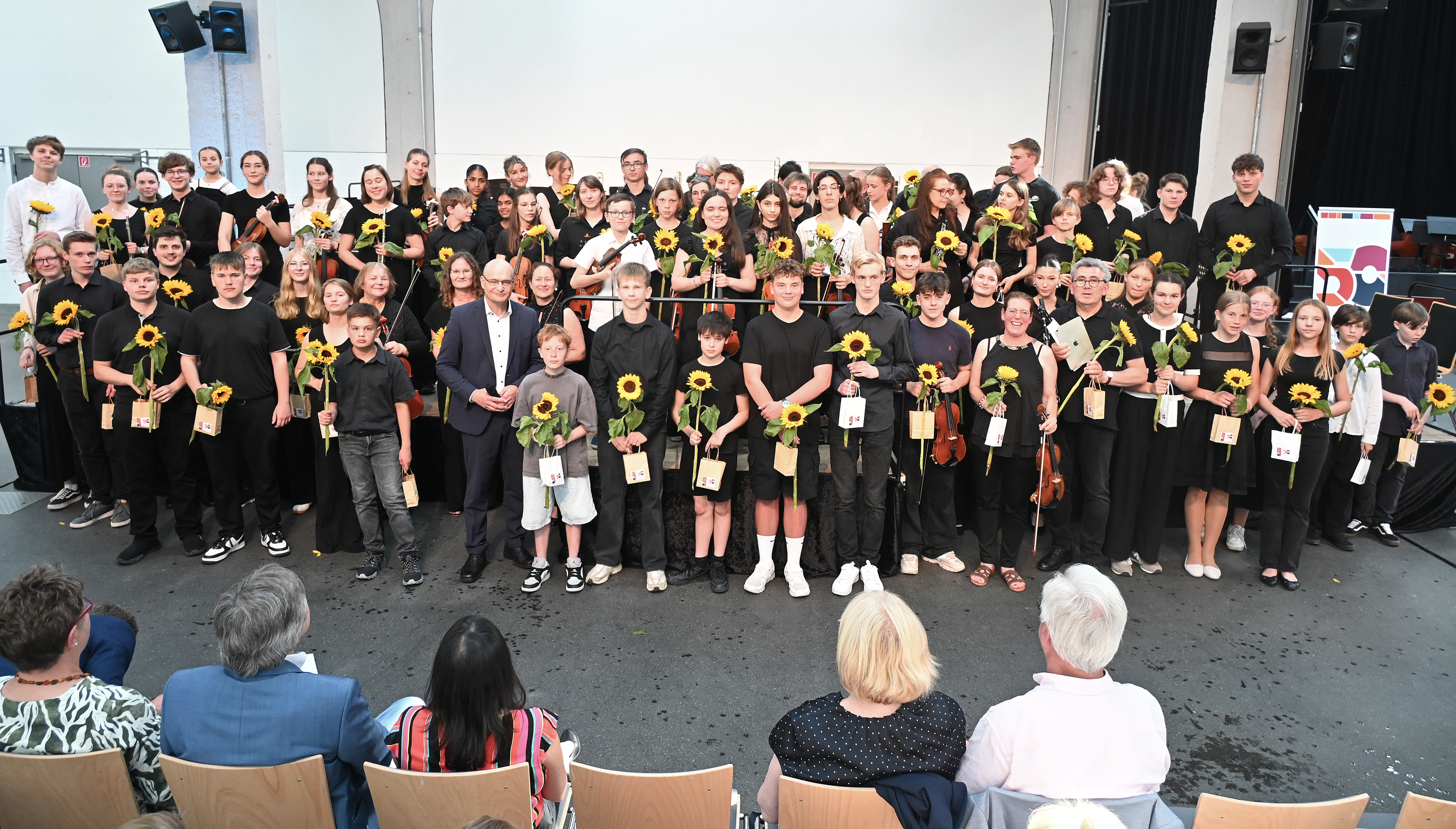 Die Musizierenden stehen auf der Bühne und davor und haben Sonnenblumen und Geschenke für ihren Auftritt bekommen.
